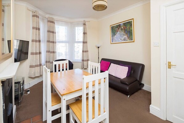 A room with a wooden dining table, white chairs, a black leather sofa, a painting on the wall, and a white door.