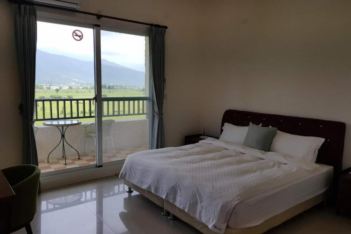 A hotel room with a bed, a small table, a chair, and a balcony with a view of the countryside.