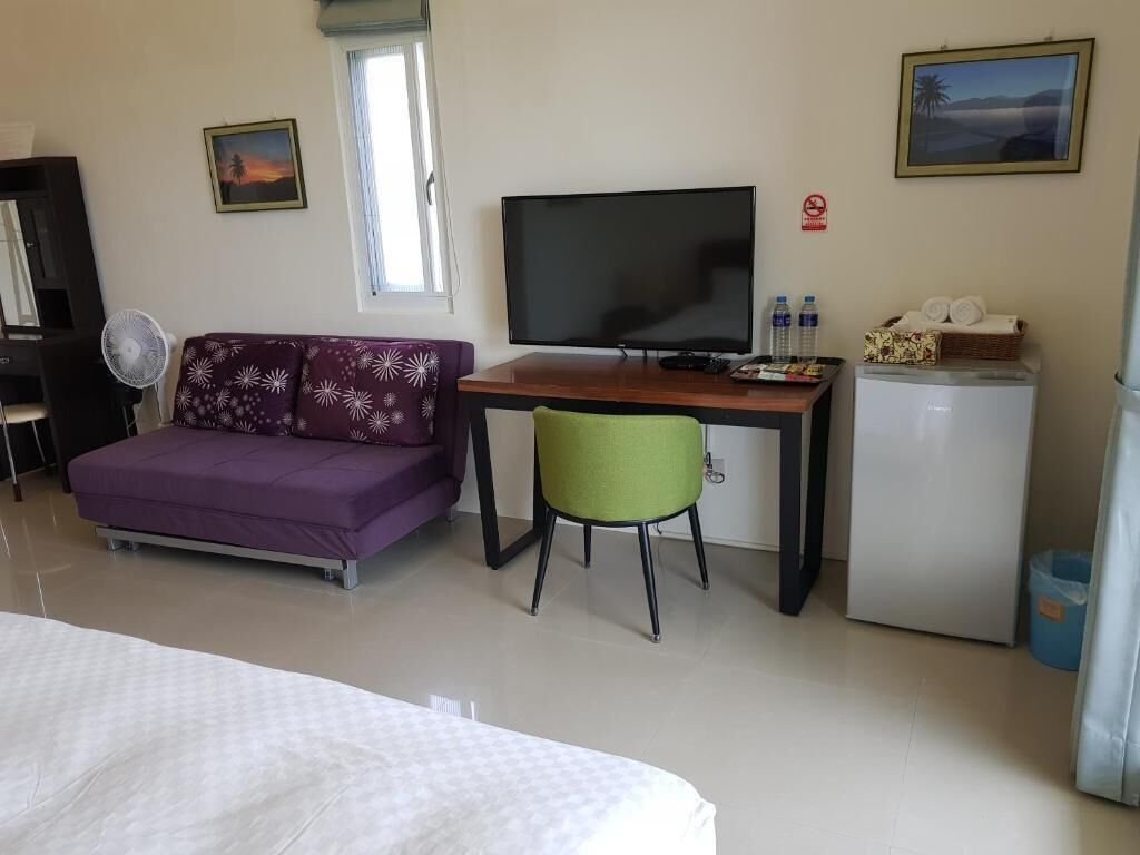 A hotel room with a bed, a purple sofa, a green chair, a TV, a small fridge, and a window with a view.
