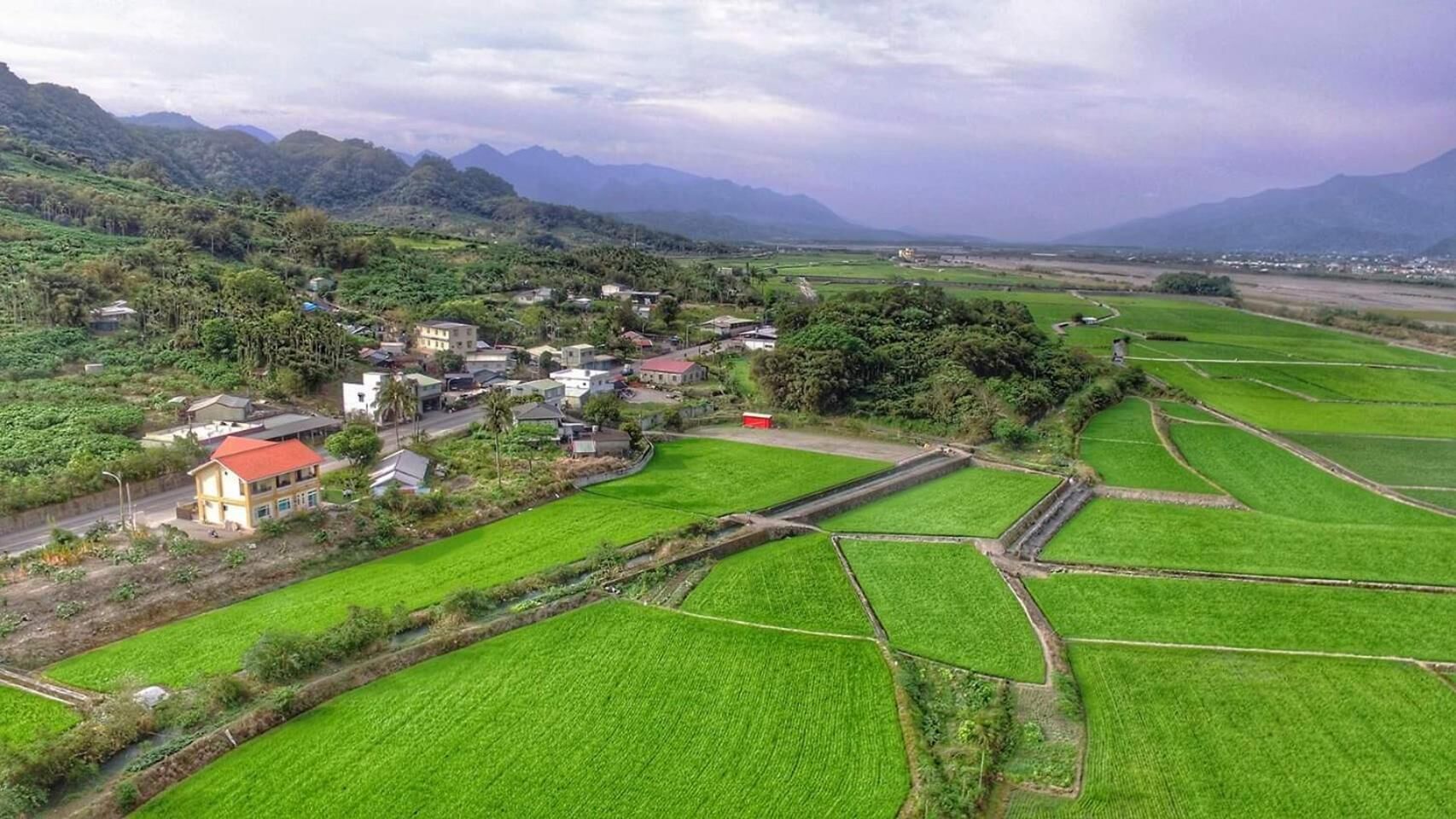 An aerial view of a rural village with terraced fields, houses, and a mountain range in the background.