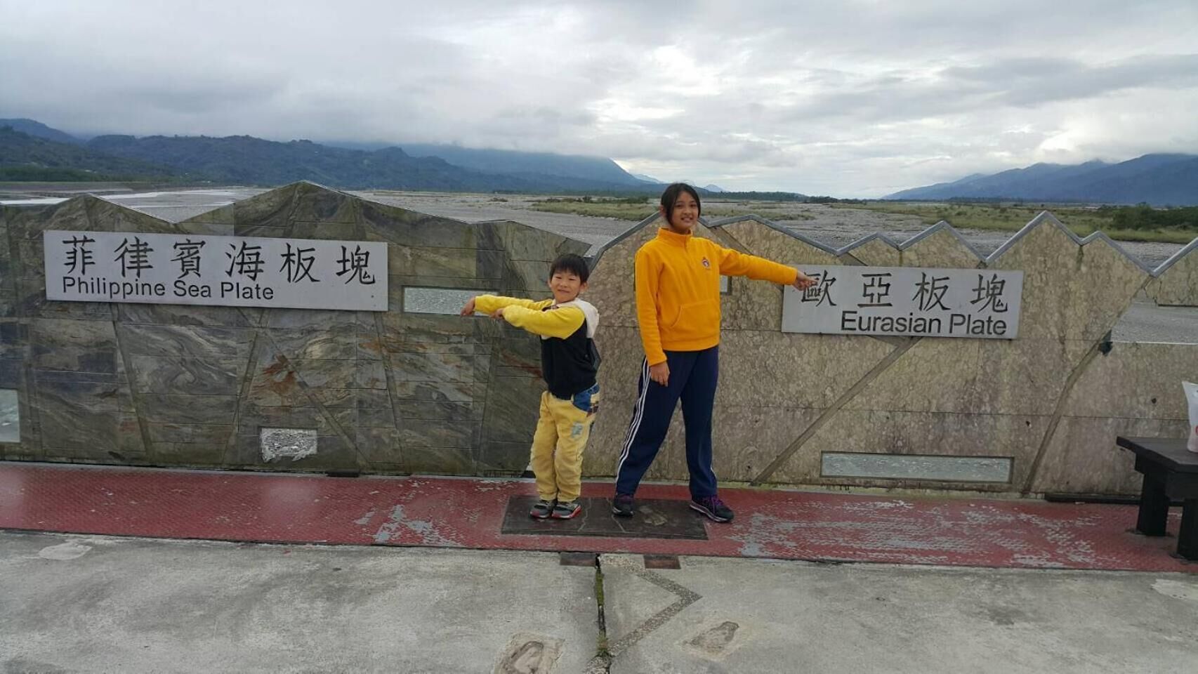 A person standing next to a signboard with text in two languages, pointing towards a landscape with mountains in the background.