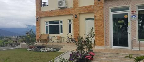 Un edificio de dos pisos con un balcón, una bicicleta estacionada afuera y un jardín bien cuidado con diversas plantas y flores.