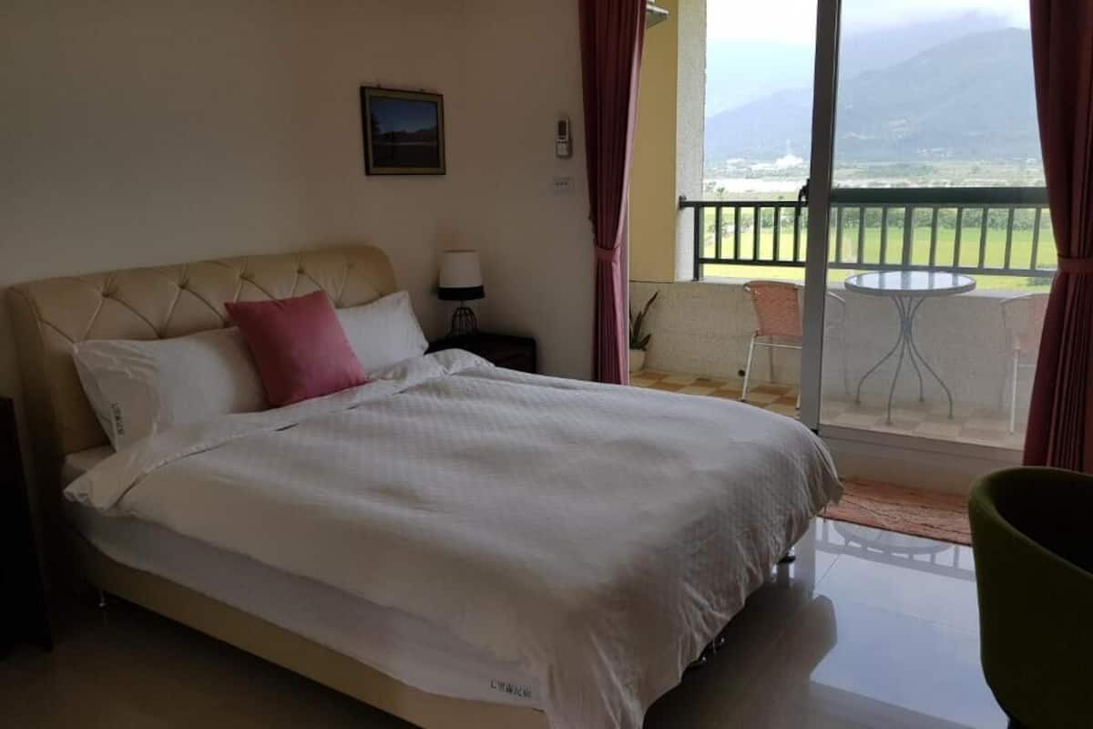 A hotel room with a bed, a nightstand, a lamp, a view of the outdoors, and a balcony with chairs.