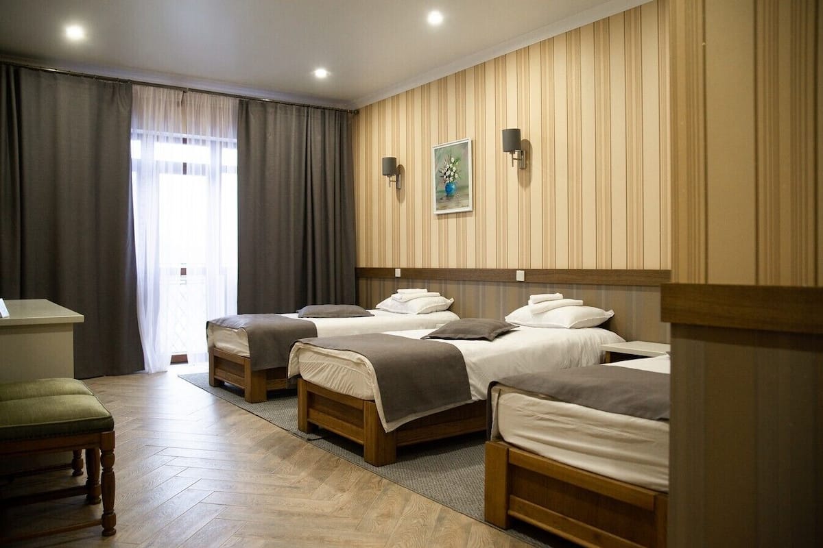 Habitación de hotel con tres camas, cabeceros de madera, un escritorio y un ventanal con cortinas.