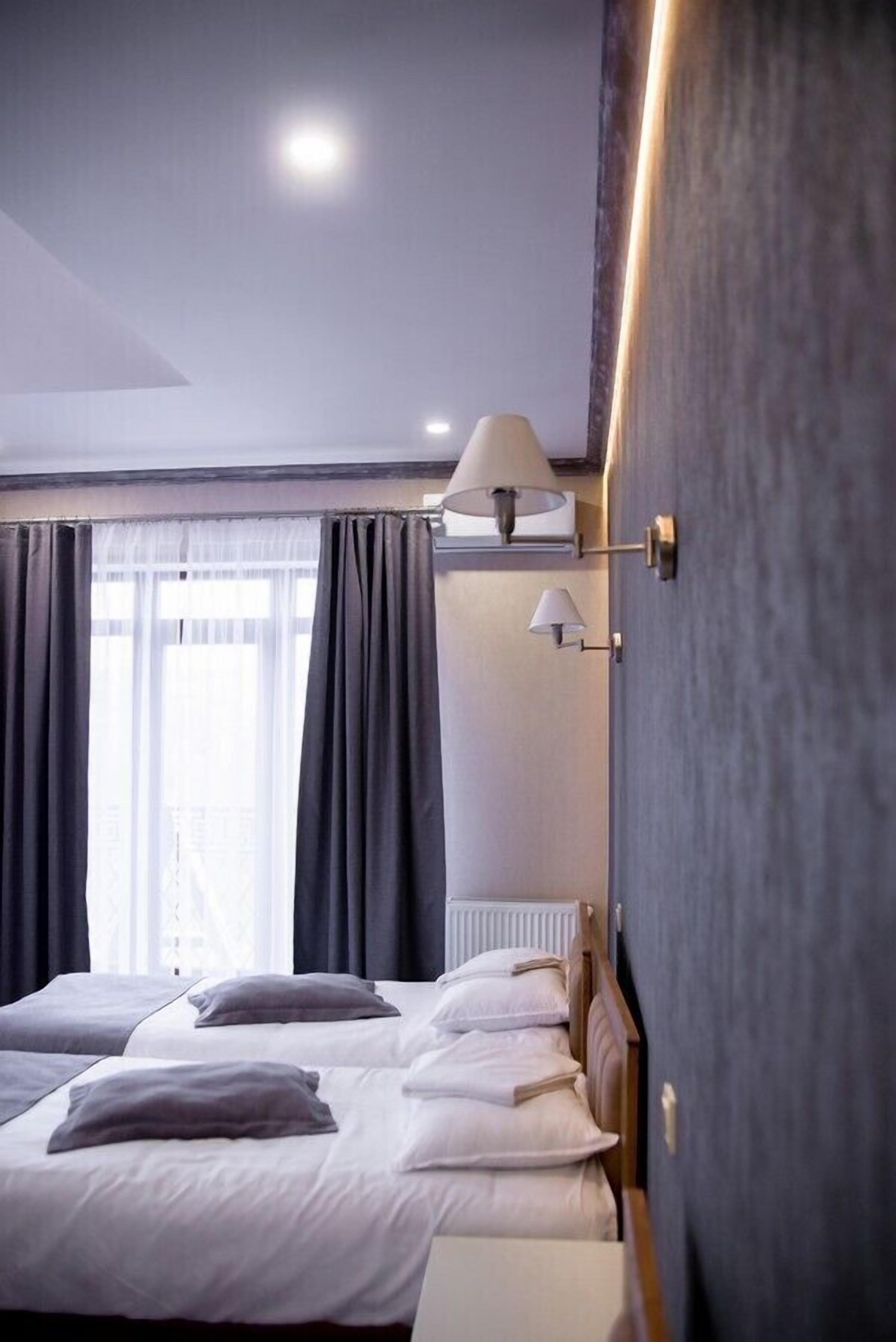 A hotel room with two beds, a window with curtains, a wall-mounted lamp, and a radiator.