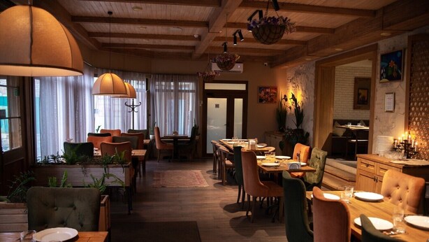 Un restaurante con vigas de madera, diferentes opciones de asientos, incluyendo cabinas y mesas, y un ambiente cálido con luces colgantes y plantas.