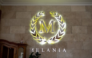 Una pared con un emblema circular que presenta una corona de laurel y la letra “M” en el centro, con la palabra “MELANIA” debajo.
