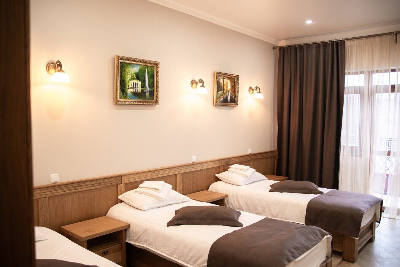 A hotel room with three beds, each with a wooden headboard, white bedding, and brown throw pillows. There are wall-mounted light fixtures and two framed pictures on the wall.