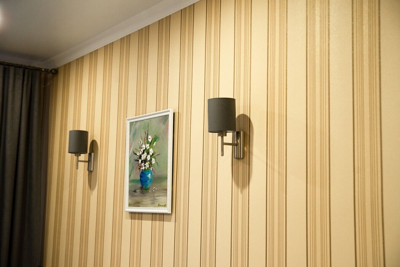 Eine Wand mit vertikalen Streifen, ein gerahmtes Bild von einer Vase mit Blumen und zwei Wandleuchten.
