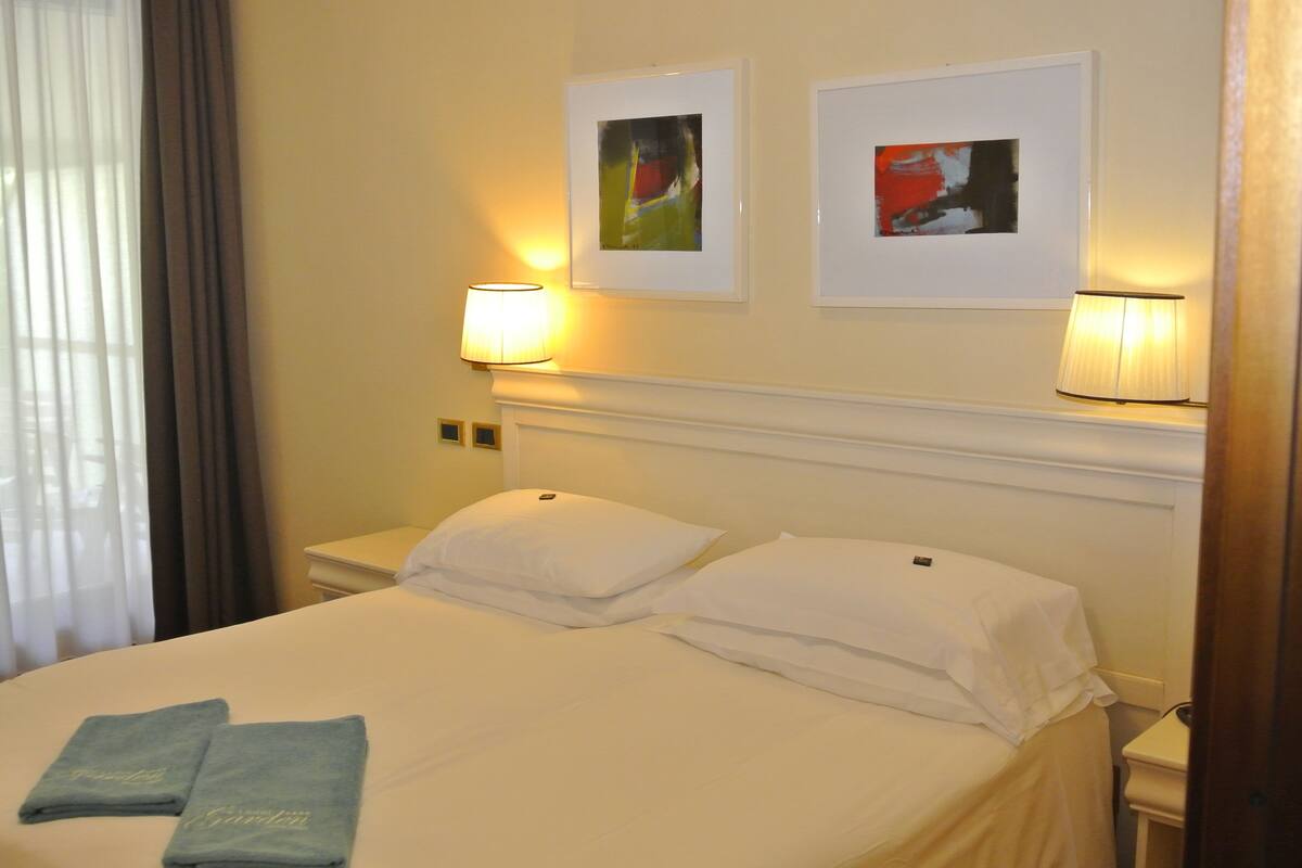 A hotel room with a bed, two pillows, two folded towels, two wall-mounted lamps, and two framed pictures.