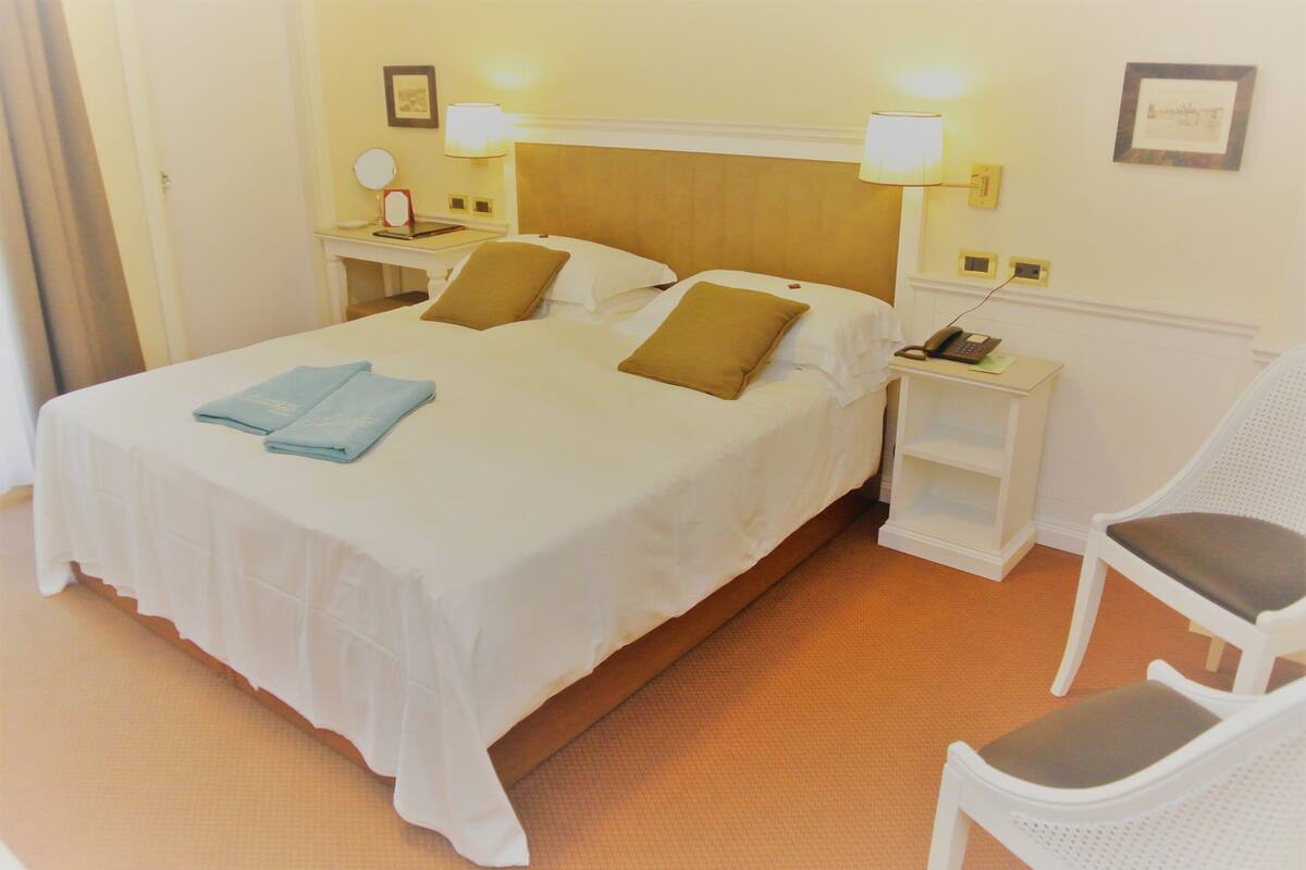 A neatly made bed with white linens and two pillows, a bedside table with a lamp, and a chair in a hotel room.