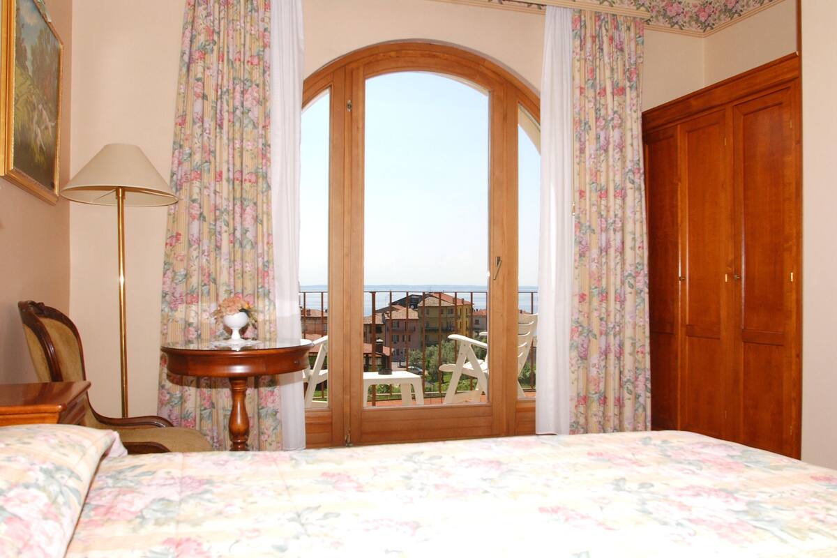 A bedroom with a bed, a chair, a table, and a balcony with a view of buildings.