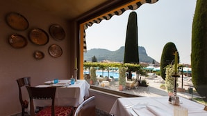 A dining area with tables set for two, a view of a pool and mountains, and a decorative wall with plates. - Hotel Garden (Garda)
