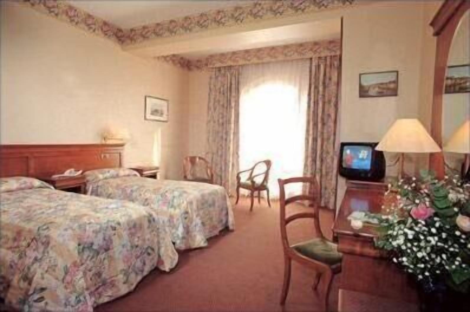 Een hotelkamer met twee bedden, een bureau met een televisie, een stoel en een raam met gordijnen.