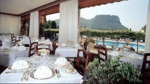 A restaurant with tables set for dining, large windows, and a view of a mountain and pool. - Hotel Garden (Garda)