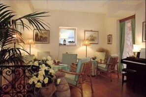 A room with a piano, chairs, a table with flowers, and framed pictures on the wall. - Hotel Garden (Garda)