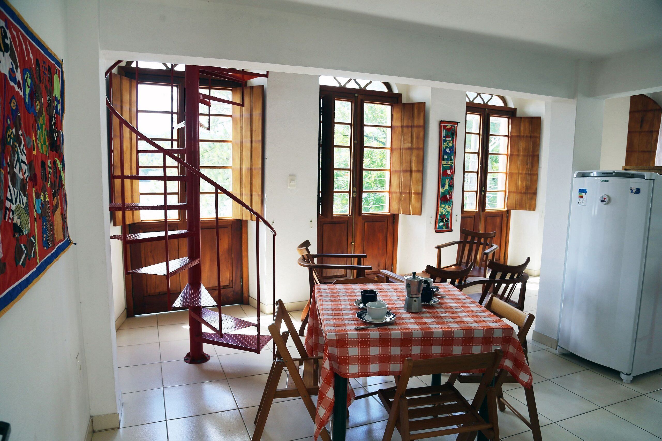 Uma área de refeições com toalha de mesa xadrez, cadeiras de madeira, escada e geladeira.