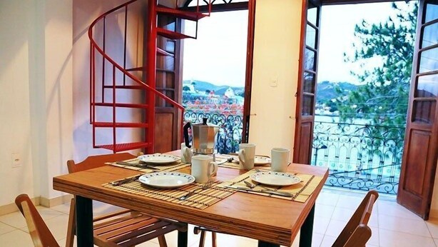 Un coin repas avec une table en bois dressée pour quatre personnes, un escalier en colimaçon et une vue sur le paysage urbain à travers des portes ouvertes.