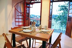 Un coin repas avec une table en bois dressée pour quatre personnes, un escalier en colimaçon et une vue sur le paysage urbain à travers des portes ouvertes.