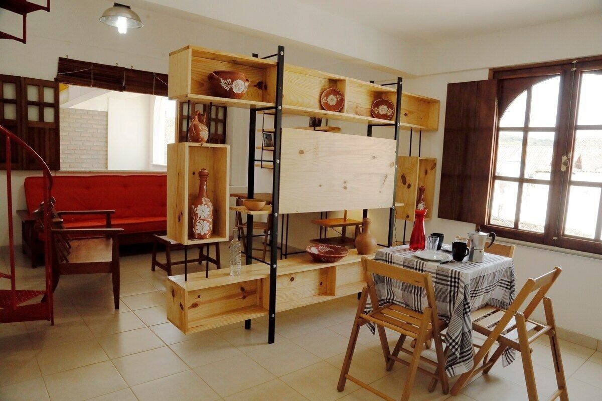A dining area with a wooden table and chairs, a red sofa, and a wooden shelf unit with dishes.