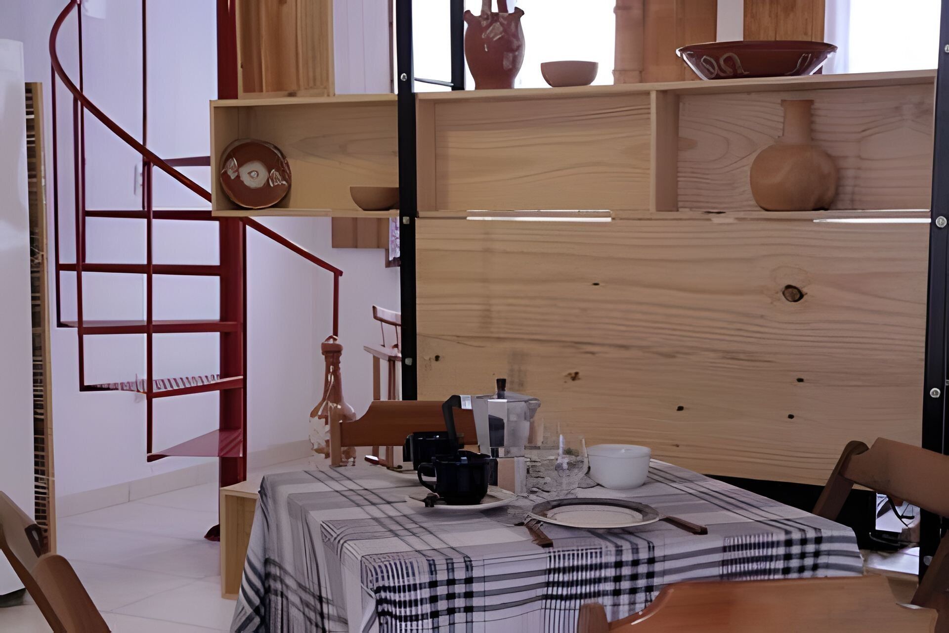 A dining area with a checkered tablecloth, wooden chairs, a red spiral staircase, and shelves with pottery and kitchenware.