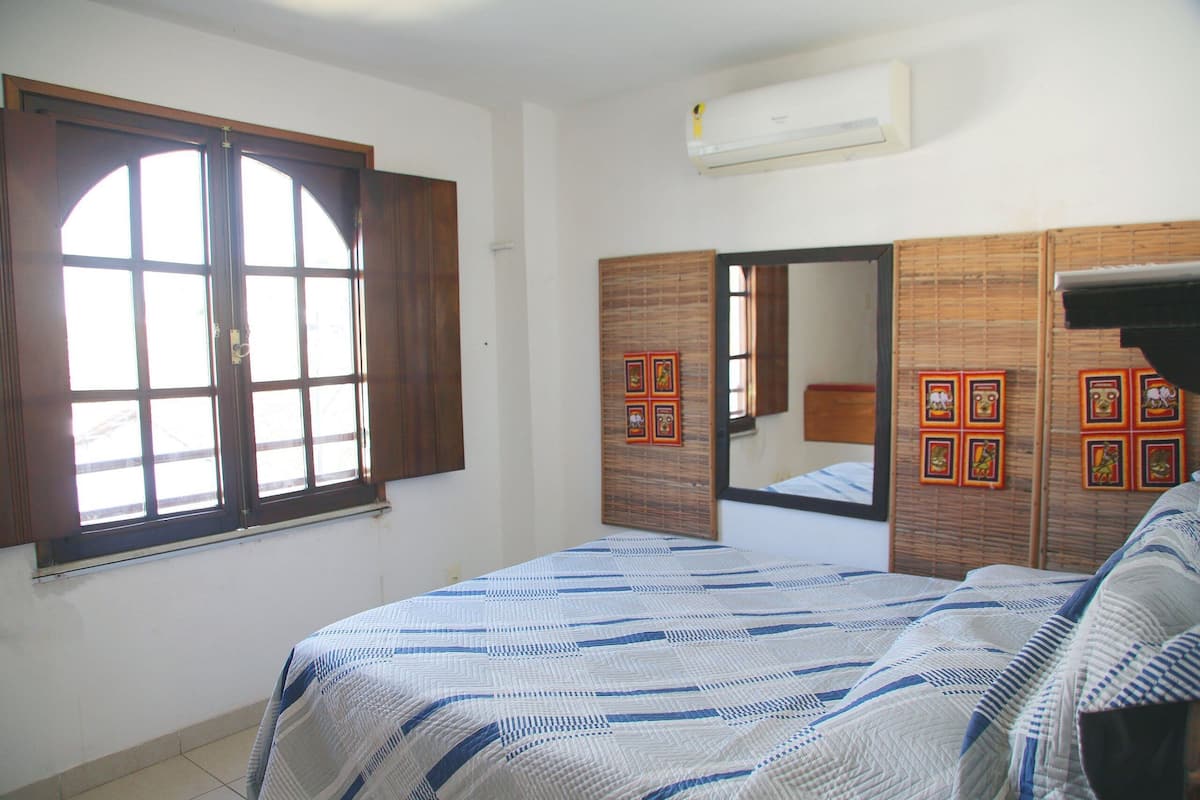 Une chambre à coucher avec un grand lit, une fenêtre avec des volets en bois, un miroir et un climatiseur fixé au mur.