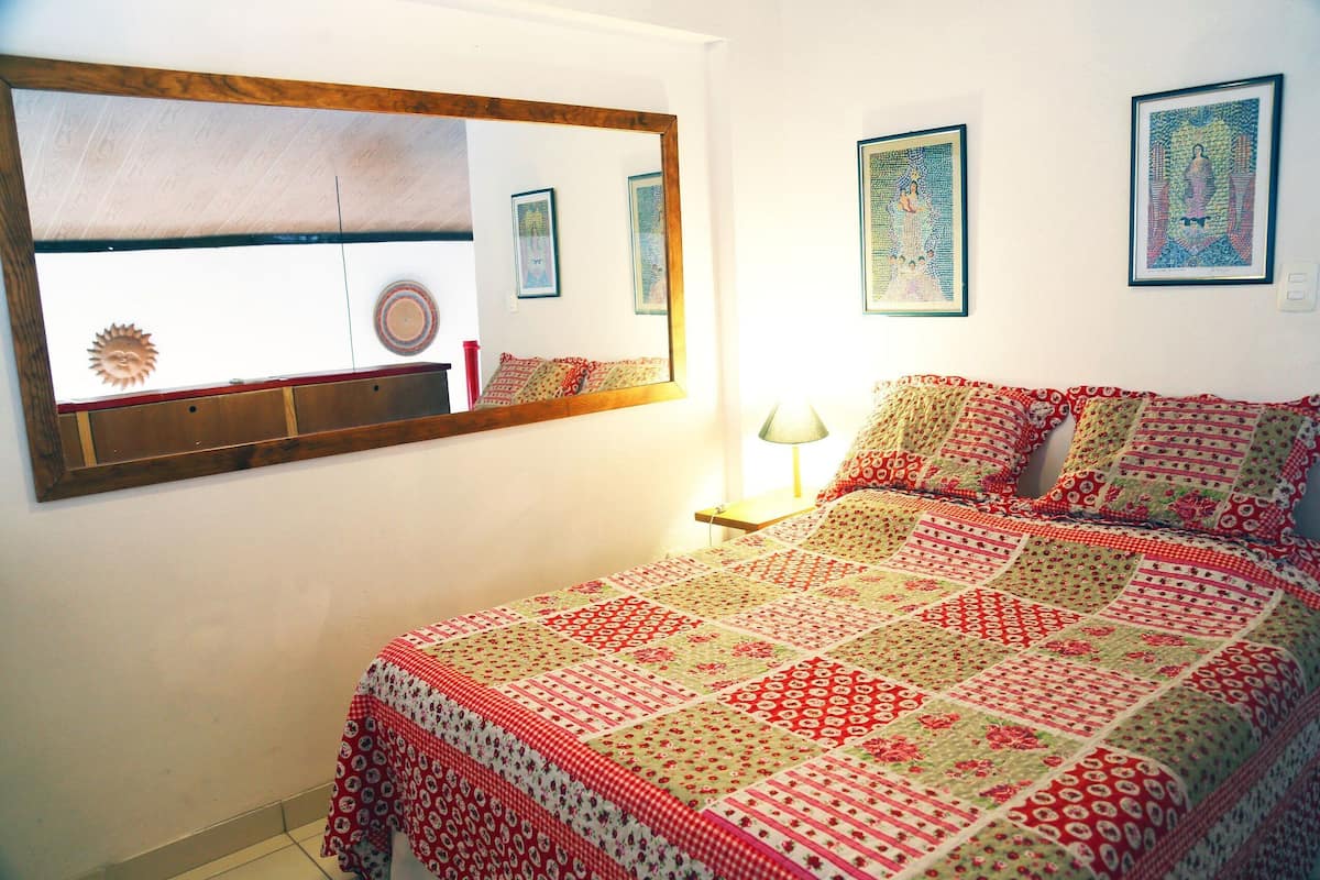 Une chambre avec un grand miroir, une couvre-lit matelassée et deux œuvres d’art encadrées accrochées au mur.