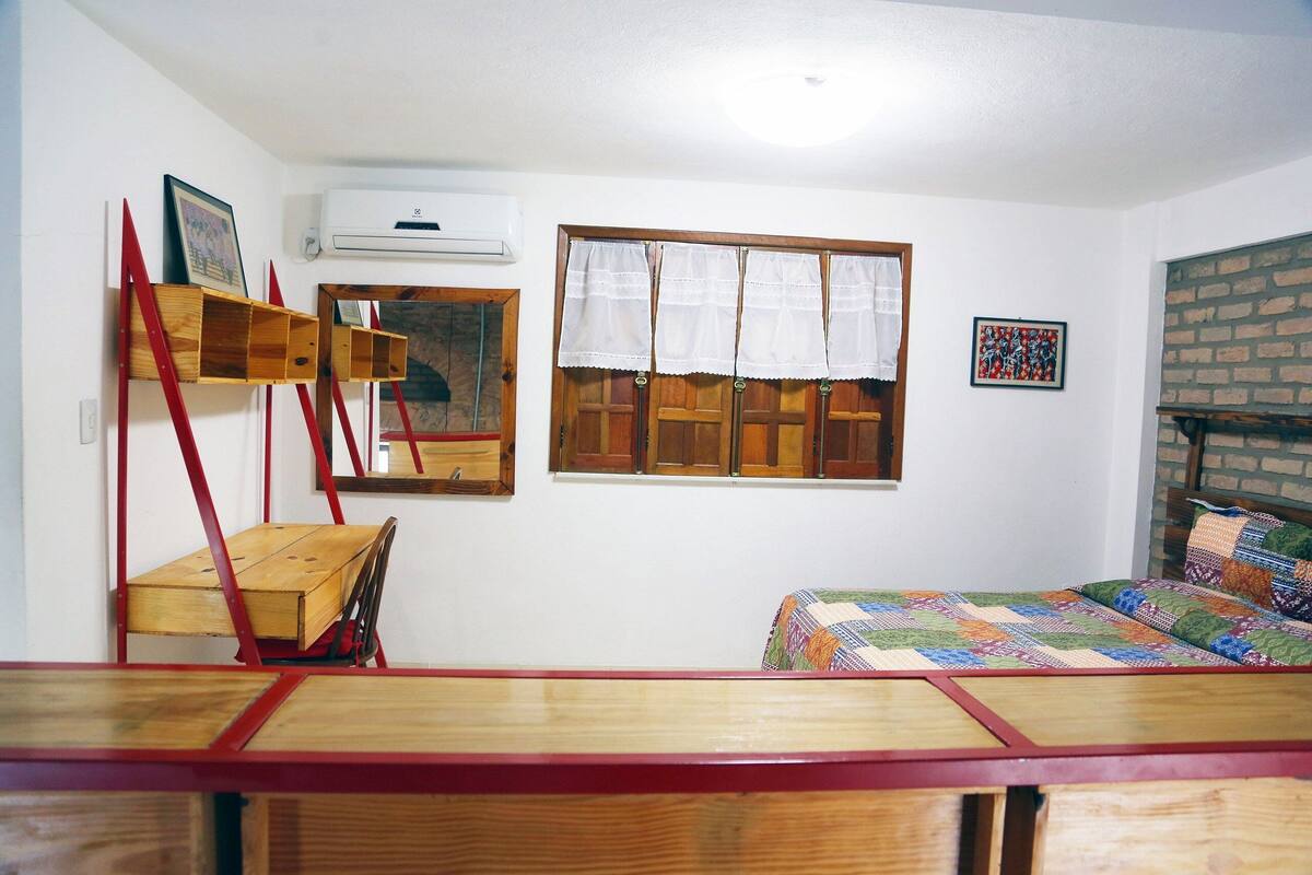Une pièce comprenant un lit en bois, un bureau en bois, une chaise, une fenêtre avec des rideaux, un miroir et un appareil de climatisation.