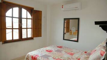 A bedroom with a bed, a window with wooden shutters, a wall-mounted air conditioner, and a mirror.