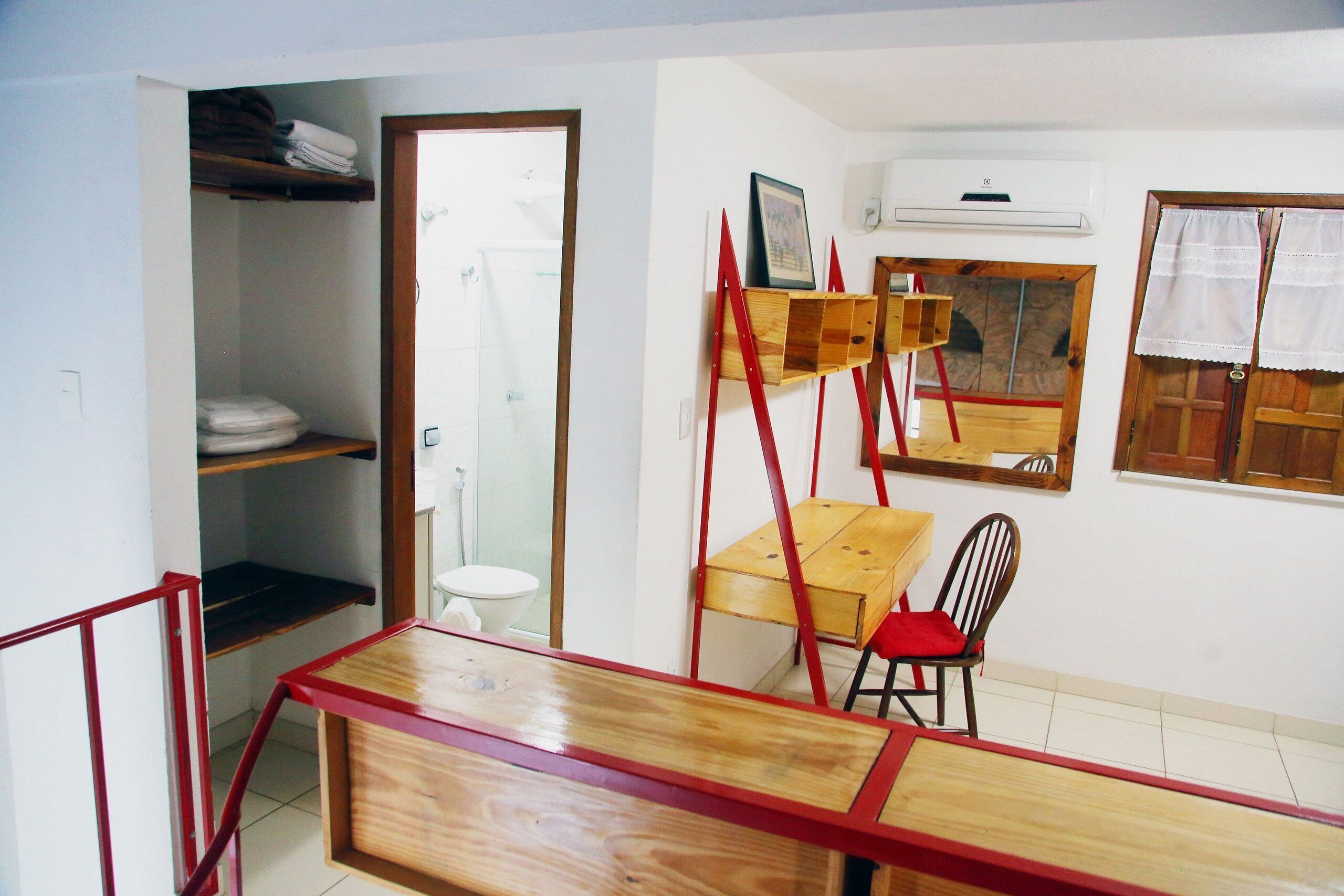 Une petite pièce avec une table en bois et une chaise, une étagère avec des serviettes pliées, des toilettes blanches et une fenêtre avec des rideaux.