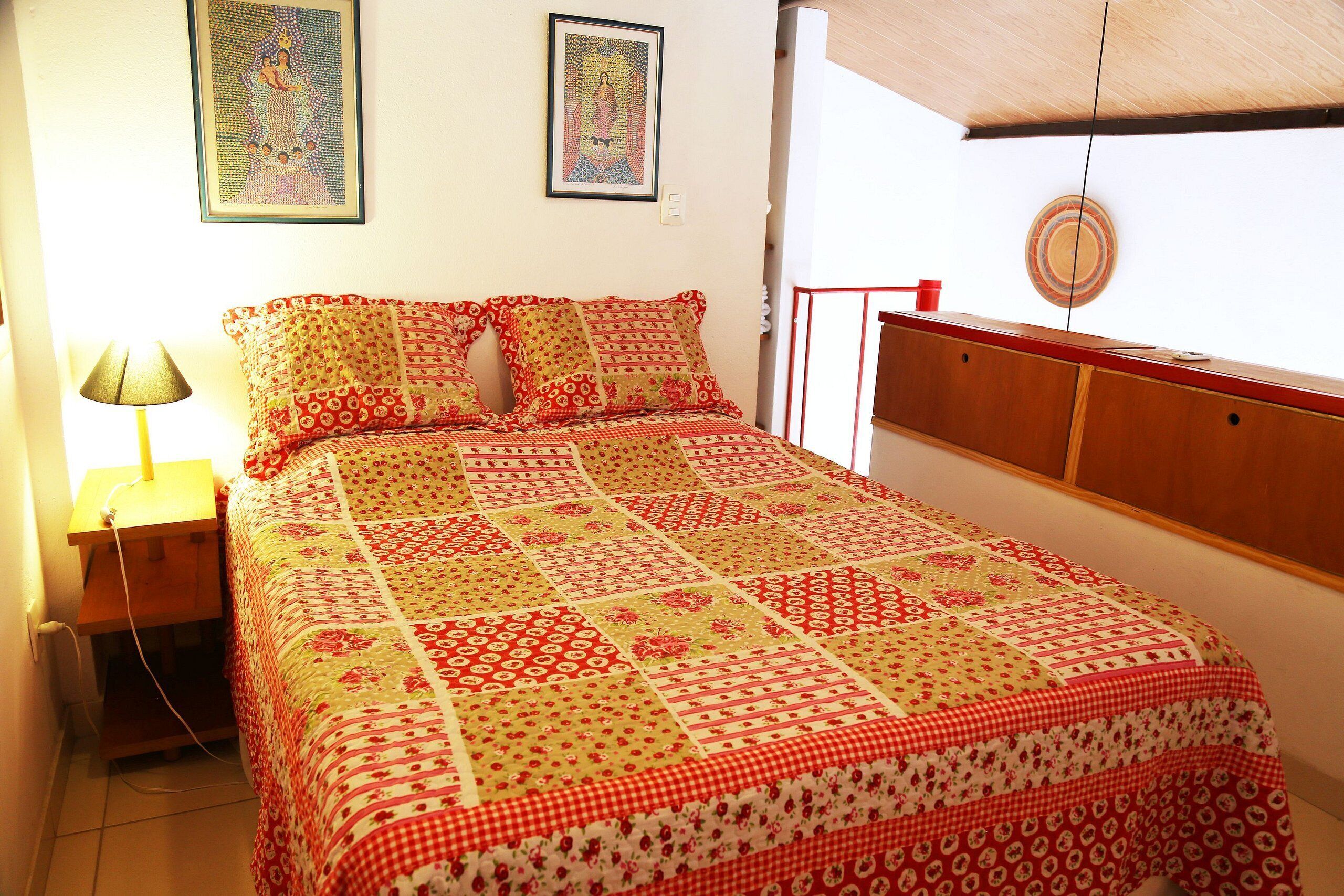 Una cama bien tendida con un edredón estampado, una lámpara de noche y dos cuadros en la pared.