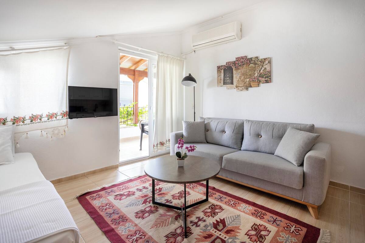 A modern living room with a grey sofa, a round coffee table with a flower vase, a flat-screen TV mounted on the wall, and a patterned rug on the floor.