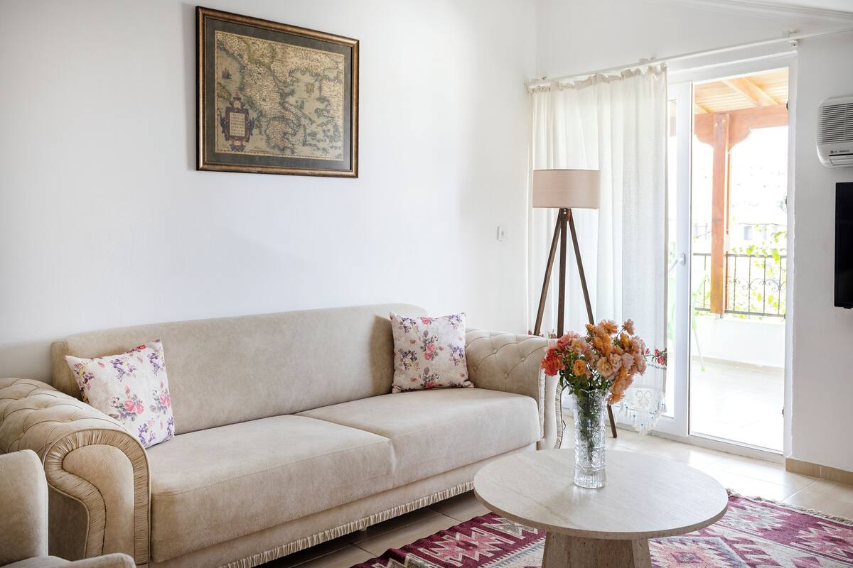 A living room with a beige sofa, a round coffee table, a vase with flowers, a framed map on the wall, and a sliding glass door leading to an outdoor area.