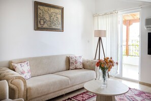 A living room with a beige sofa, a round coffee table, a vase with flowers, a framed map on the wall, and a sliding glass door leading to an outdoor area. - Kaya Apart Datca (Datça)