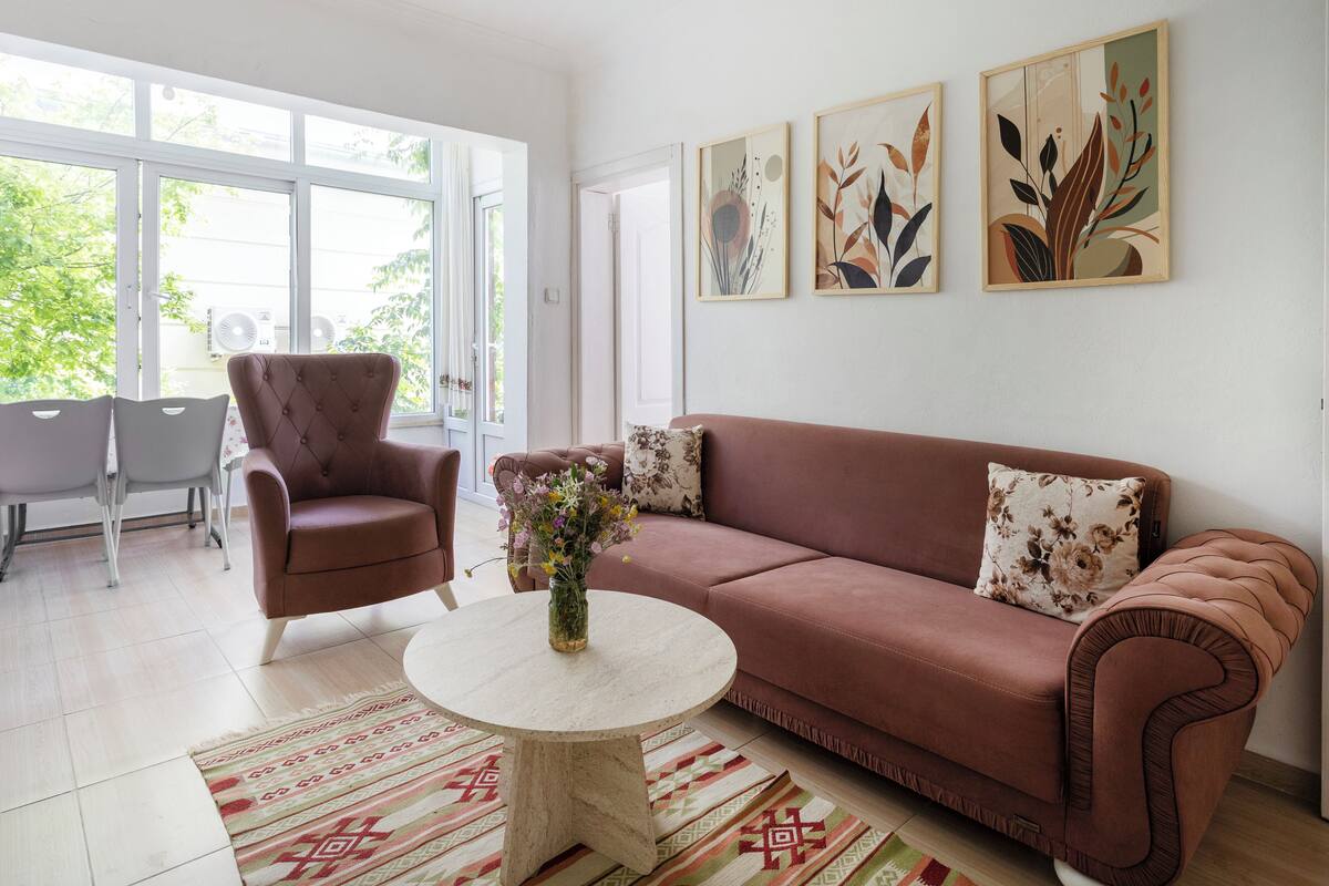 A living room with a brown sofa, a round coffee table, and a floral vase. There are two chairs and a dining table in the background.