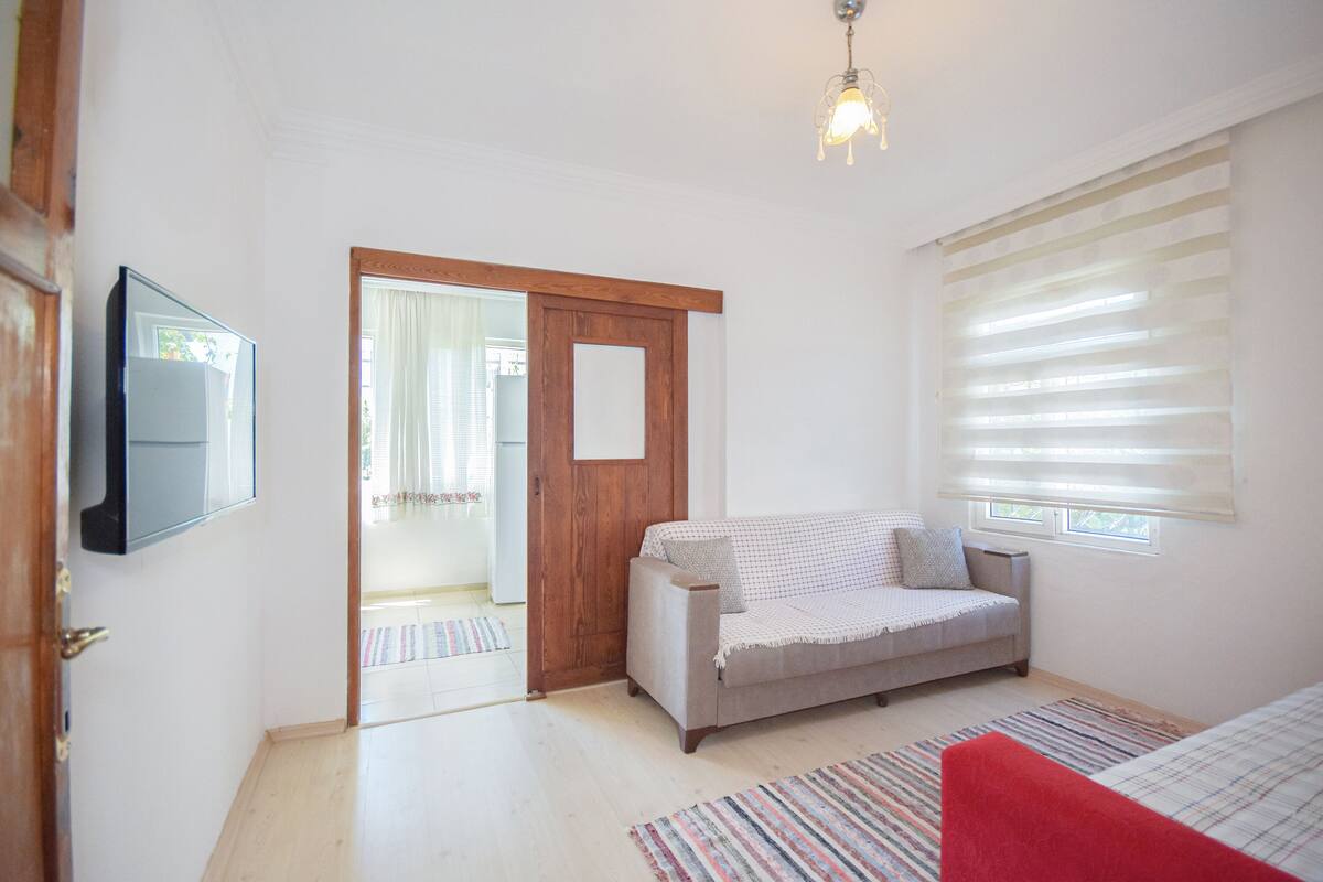 A room with a wooden door, a sofa, a TV mounted on the wall, and a window with blinds.