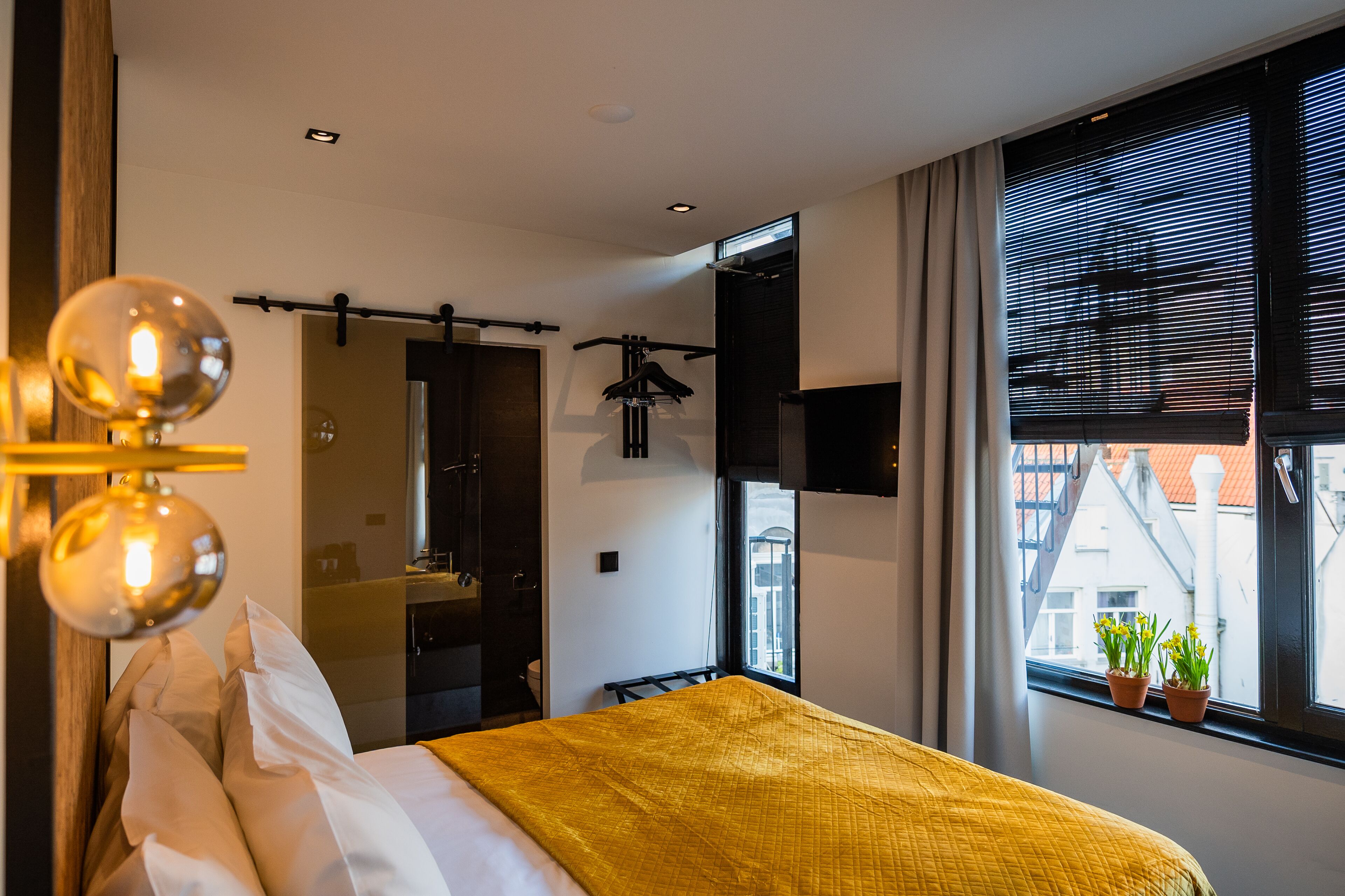 Una habitación de hotel moderna con cama, lámpara colgante, ventana con persianas y vista a edificios.
