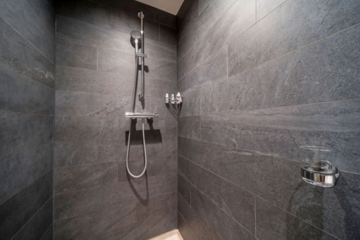 A shower with a handheld showerhead and a wall-mounted control panel, set in a room with dark tiled walls.