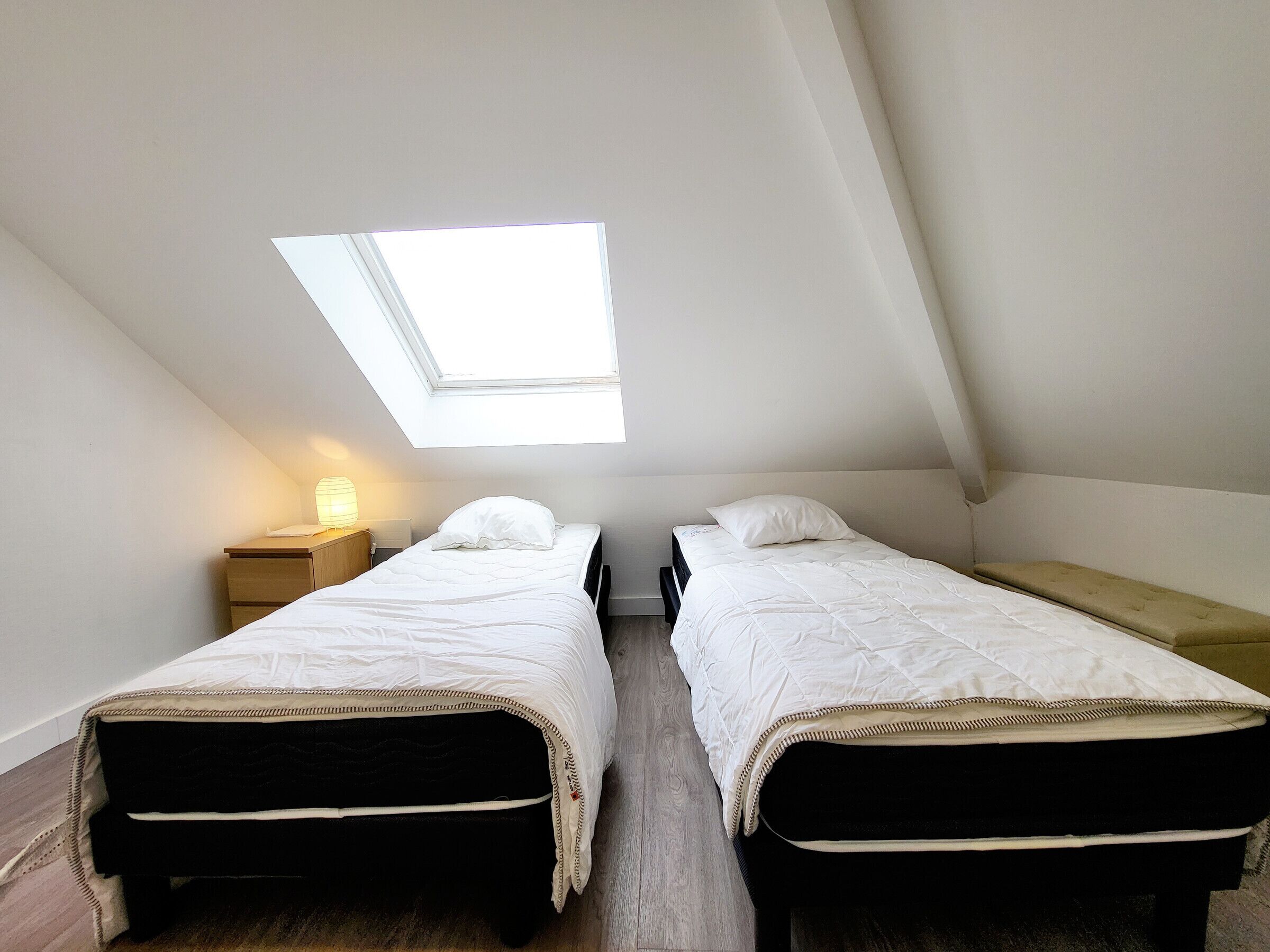 Een kleine, zwak verlichte kamer met twee eenpersoonsbedden, een dakraam en een nachtkastje met een lamp.