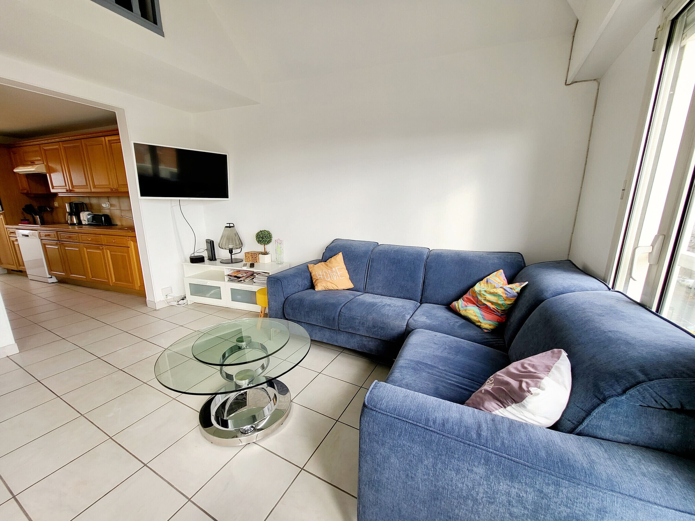A living room with a blue sofa, a glass coffee table, and a kitchen area visible in the background.