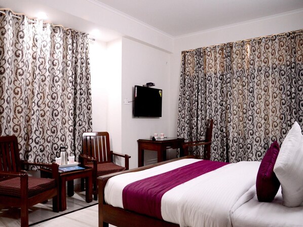 A hotel room with a bed, two wooden chairs, a small table, a flat-screen TV mounted on the wall, and patterned curtains.