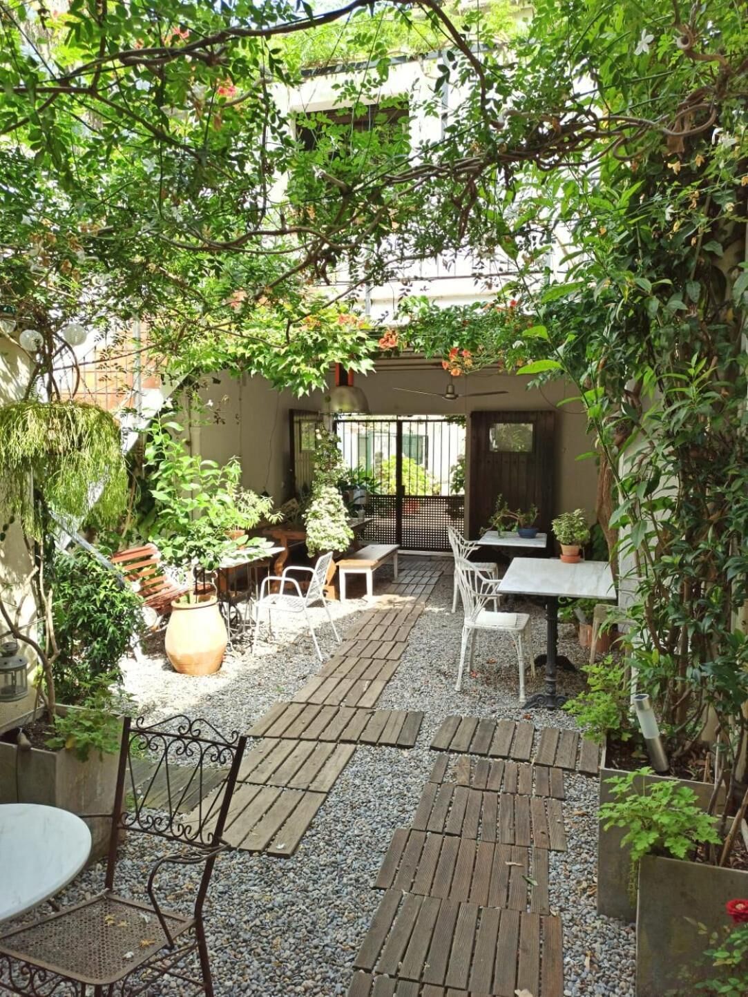 Eine Terrasse im Freien mit Holzboden, weißen Metallstühlen und -tischen. Übertöpfe mit Pflanzen und ein kleiner Gartenbereich sind ebenfalls vorhanden.