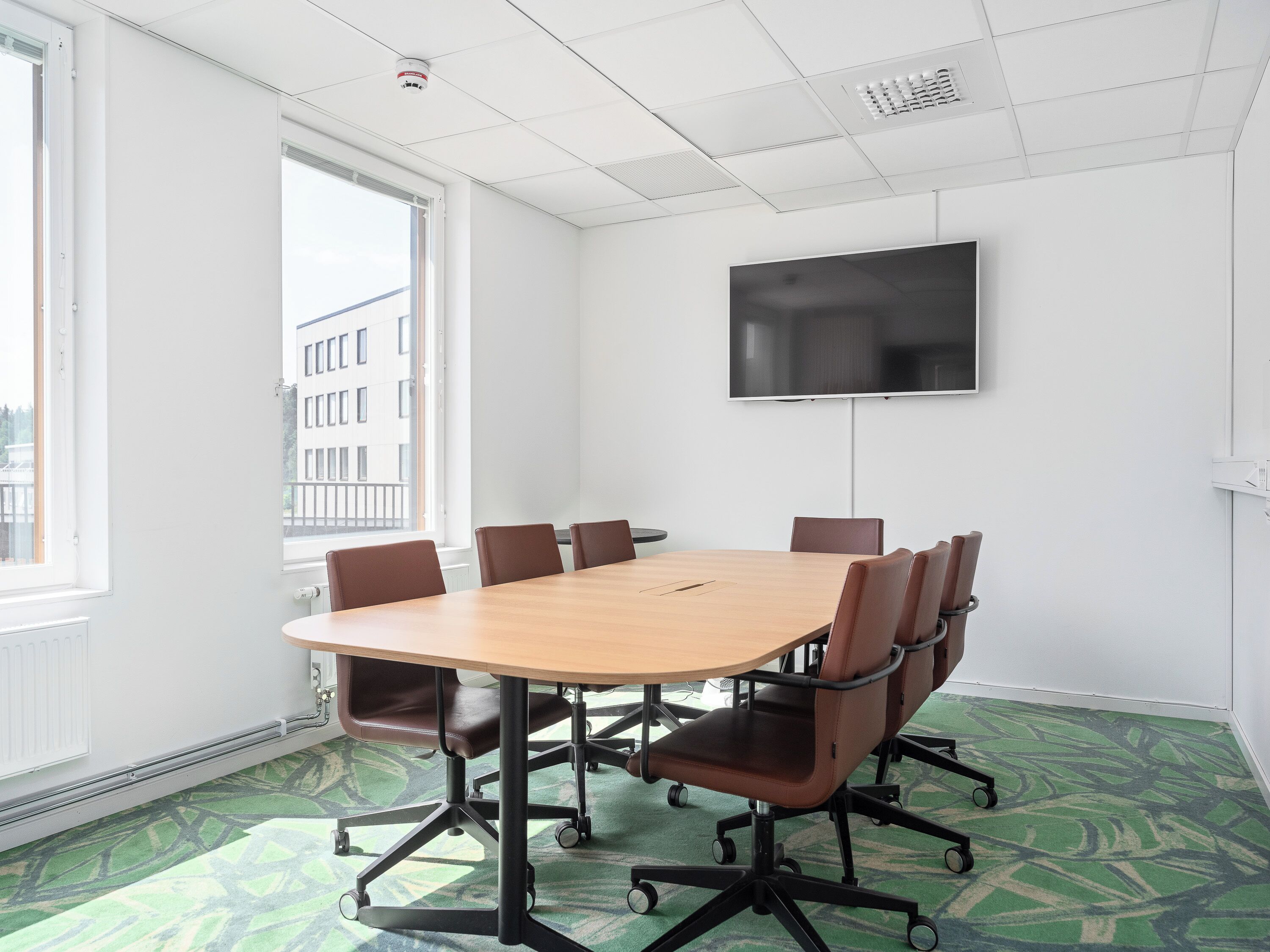 A conference room with a large table, six chairs, a flat-screen TV, and a window with a view of buildings.