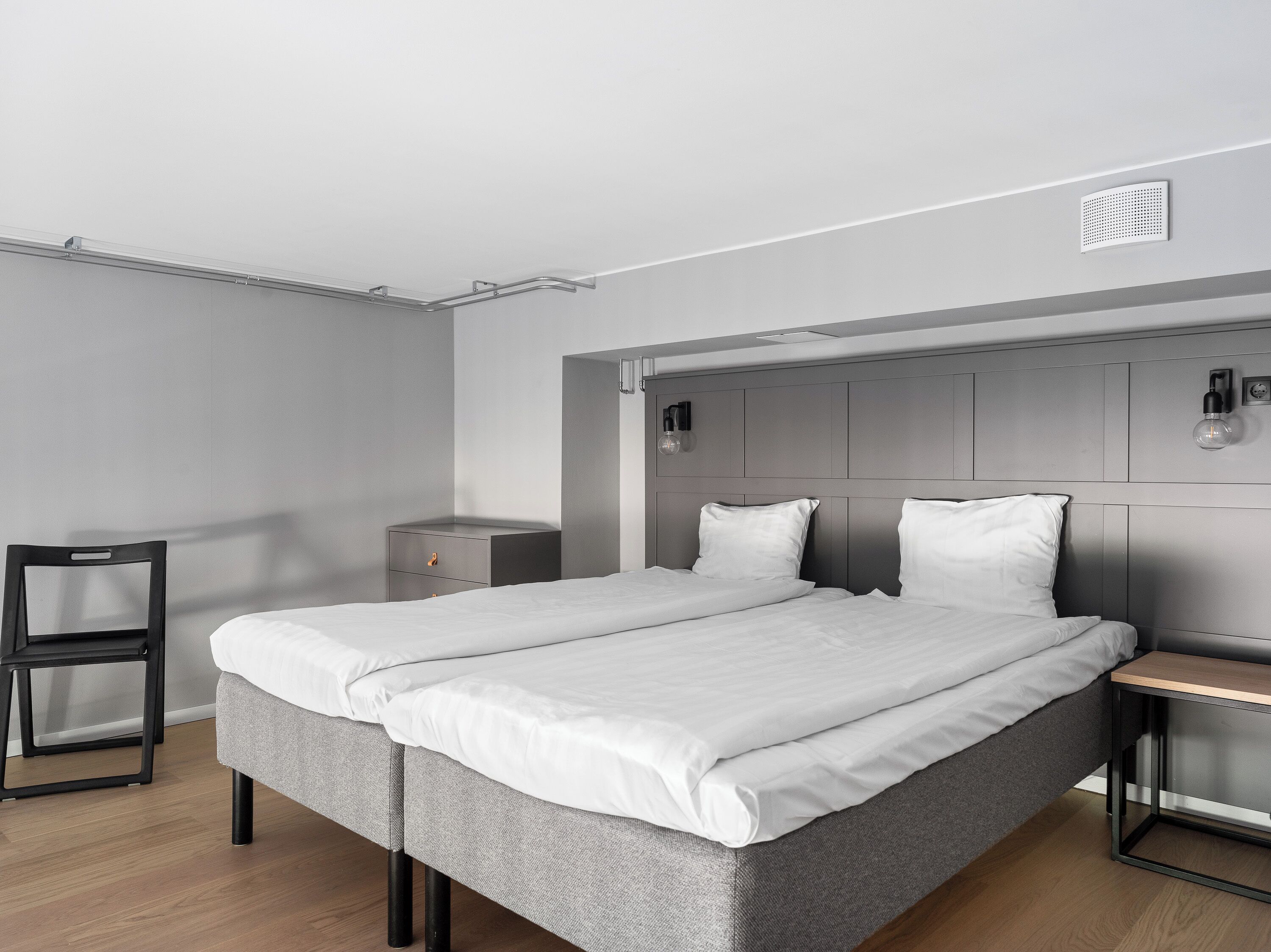 Un dormitorio moderno con una cama tapizada en gris, una mesita de noche pequeña, una silla y una lámpara fijada a la pared.