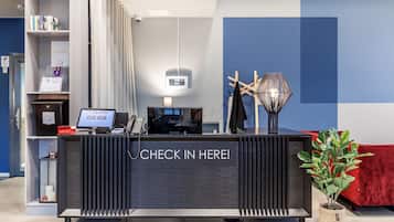 Una mesa de recepción con un letrero que dice 'CHECK IN HERE!', una computadora, un teléfono y una planta en maceta.