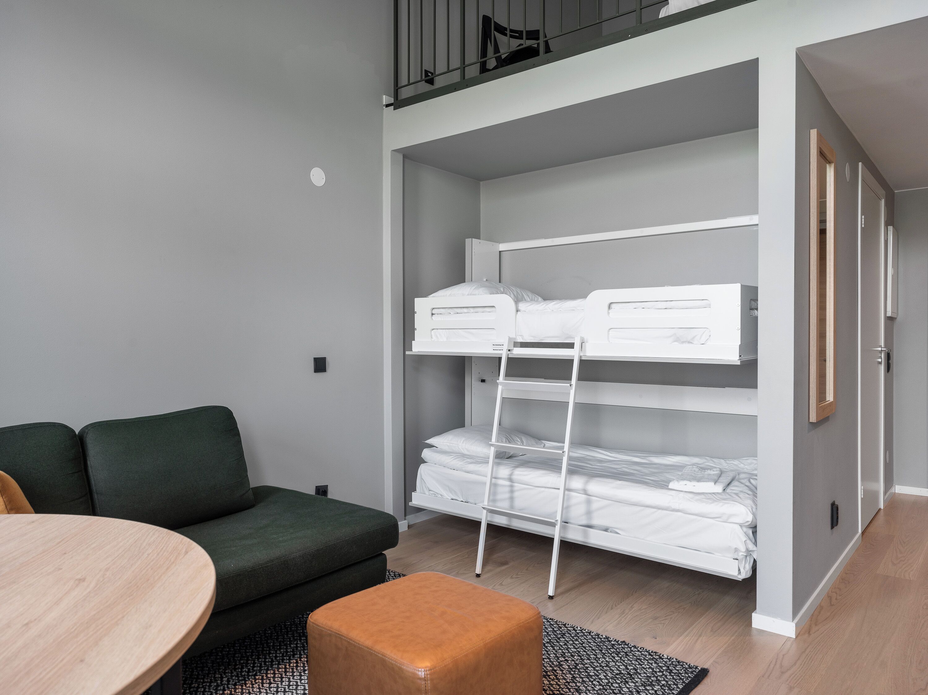 A compact room with a bunk bed, a green sofa, a wooden table, and a small orange ottoman.