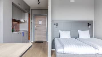 A modern hotel room with a bed, bedside tables, a built-in kitchen with a sink, and a door leading to another room.