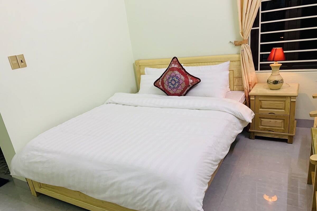 Un lit bien fait, avec du linge de lit blanc, une tête de lit en bois, un coussin décoratif, une table de chevet avec une lampe et une fenêtre avec des rideaux.