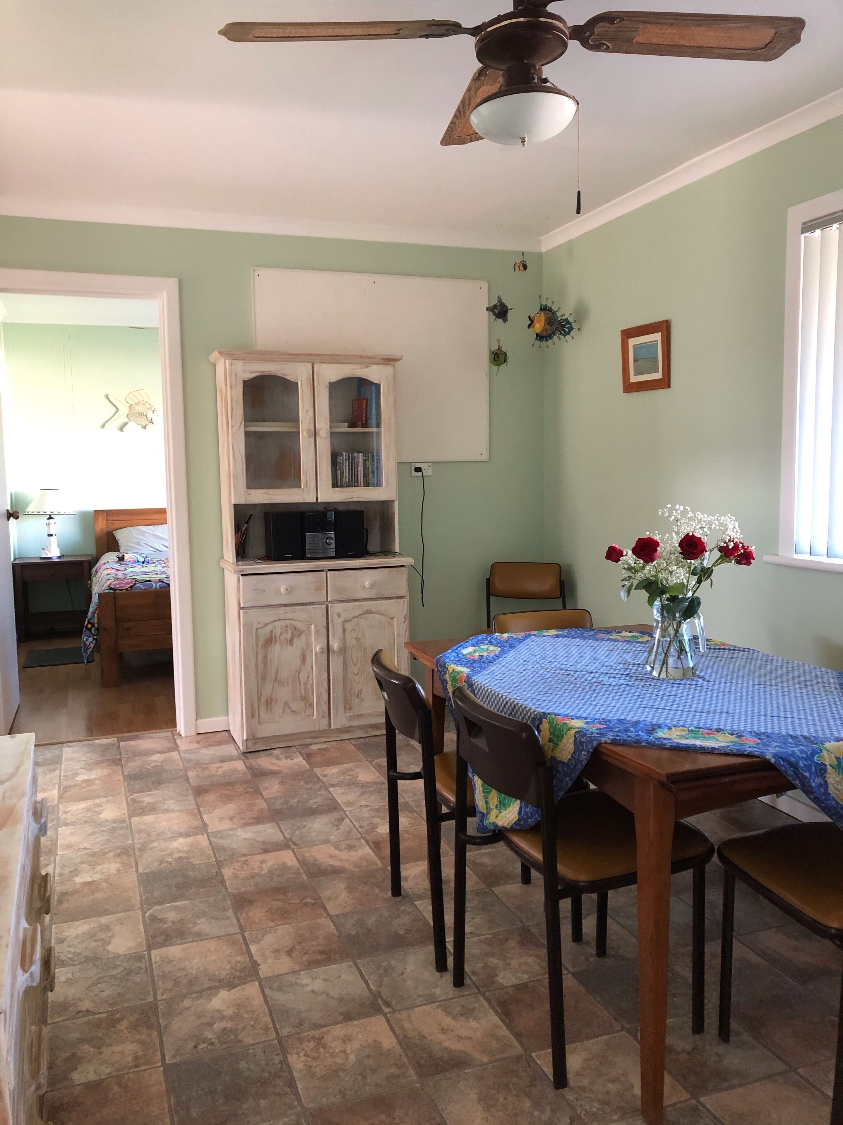 A dining area with a table set for four, a ceiling fan, a cabinet with books, and a window with blinds.