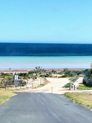 A coastal road with a speed limit sign, leading to a beach area with sparse vegetation and a clear view of the ocean. - Cosy home, within walking distance to north beach. (North Beach)